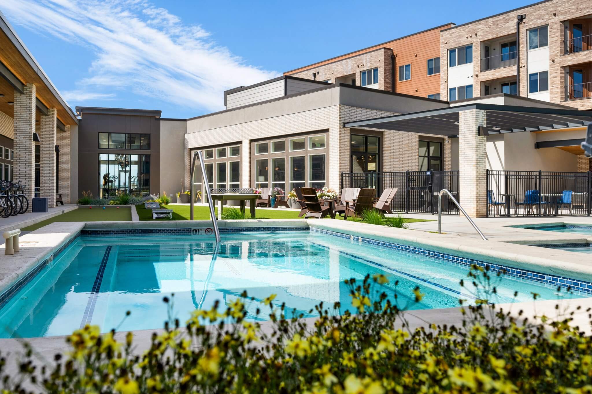 Swimming pool with chaise lounge seating, tanning shelf, ping pong and conversational seating with a leasing office building in the background